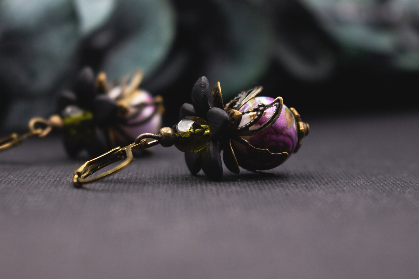 Glasperlenohrringe Lila-Oliv mit marmorierter Perle und schwarzen Blüten – ausdrucksstarker, handgefertigter Ohrschmuck im floralen Stil.