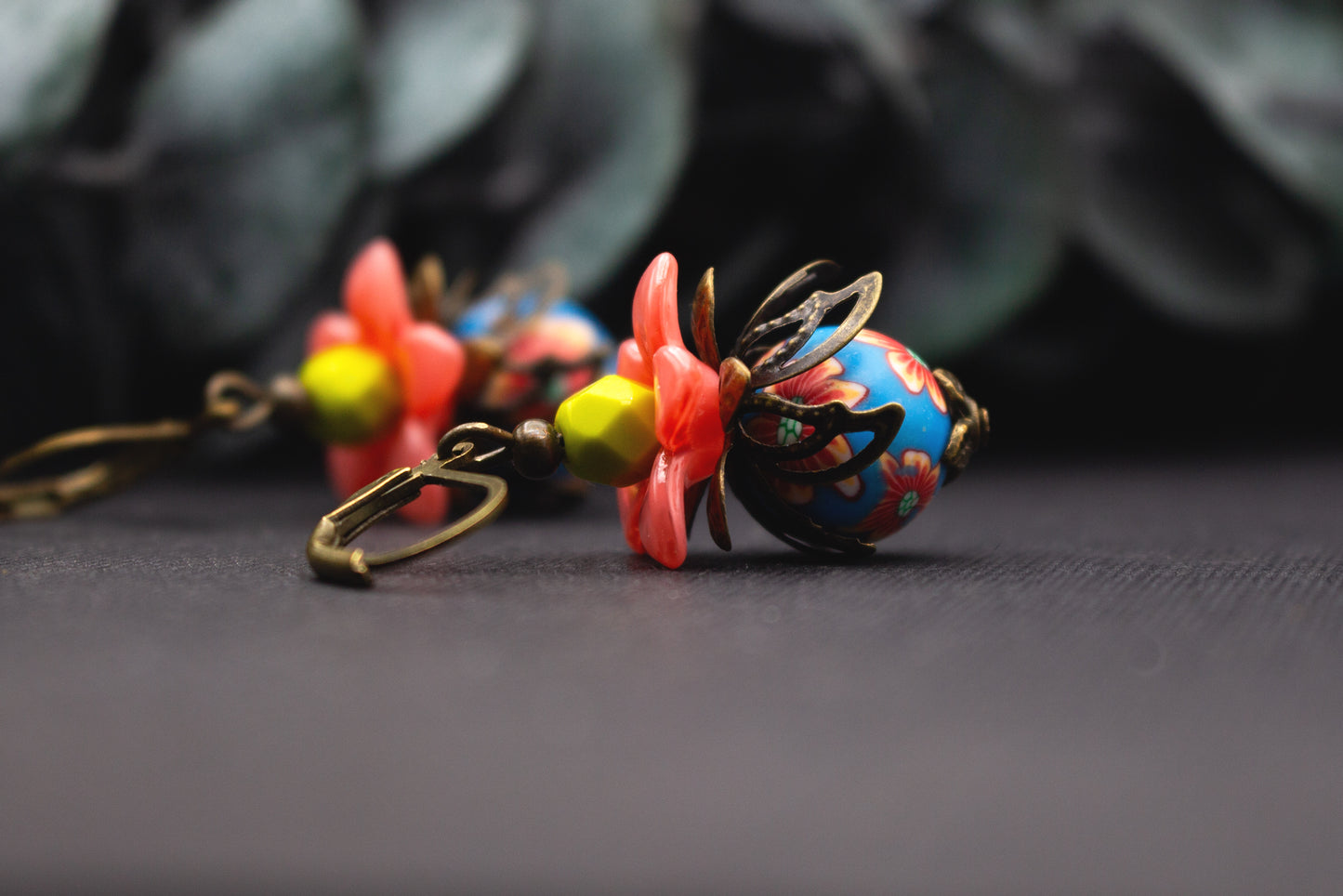 Florale Ohrringe mit lachsrosa Acrylblüte, blauer Glasperle mit roten und orangen Blumen und gelbem Mittelteil auf dunklem Hintergrund.