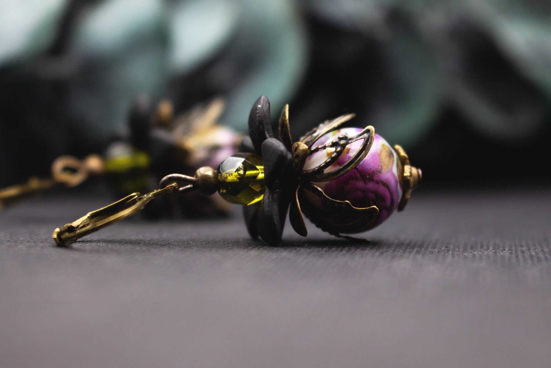 Lila-Oliv Blütenohrringe mit marmorierter Glasperle, schwarzen Acrylblüten und bronzenen Metallblättern – detailreicher Vintage-Ohrschmuck mit ausdrucksstarkem Farbspiel.