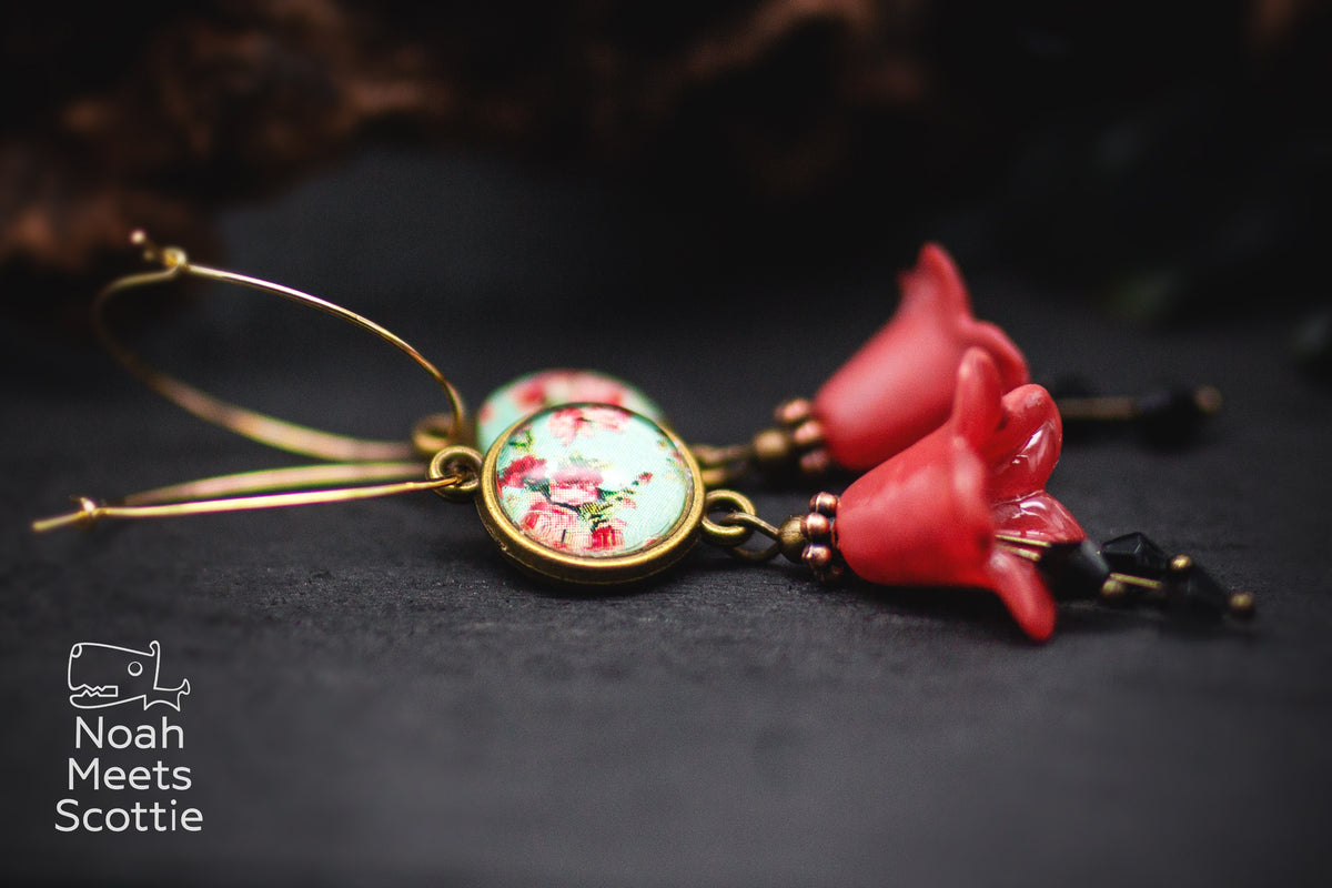 Florale Vintage Ohrringe mit roten Blüten, goldfarbenen Creolen und rundem Cabochon mit roten Rosen auf hellblauem Hintergrund.