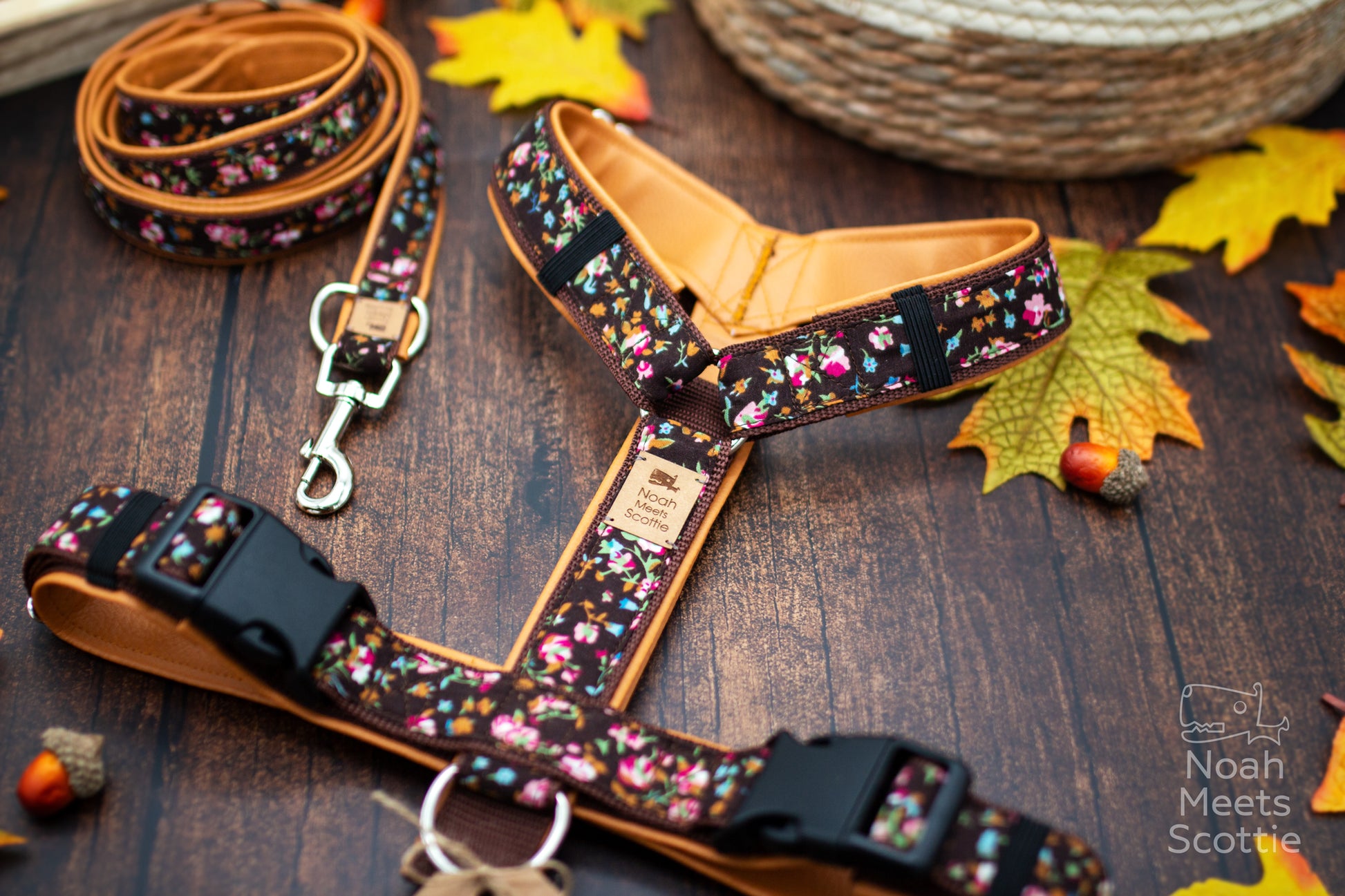 Handgemachtes Hundegeschirr Mary mit buntem Streublümchen-Muster auf dunkelbraunem Gurtband und passender Leine auf einem Holzbintergrund mit herbstlichen Blättern.